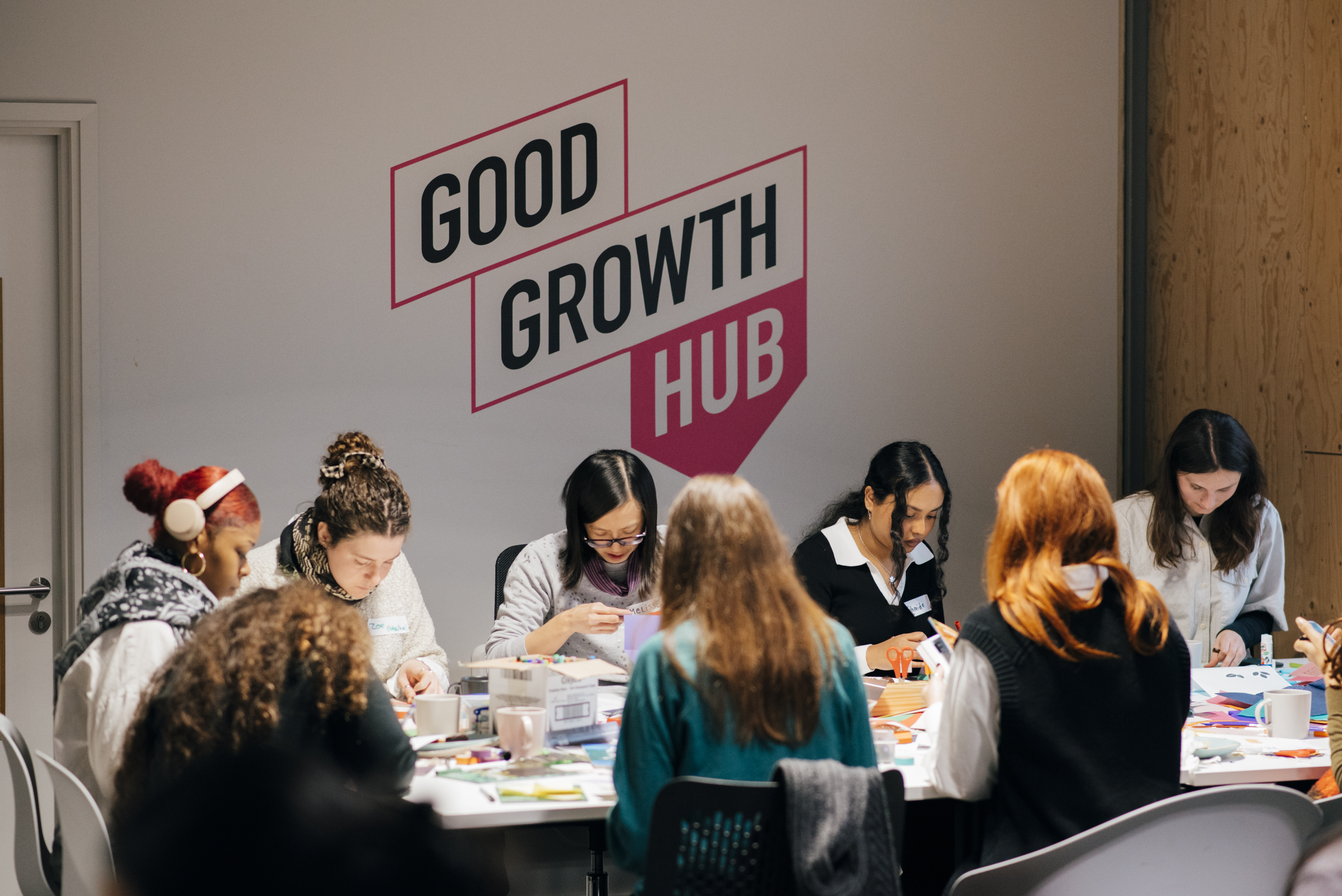 A group of people, sat in the Good Growth Hub. They are sat around a table with some people facing the camera, taking part in a zine making workshop. Behind the table on the wall is the Good Growth Hub logo.